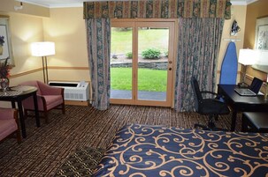 Habitación de hotel con cama, escritorio, silla, mesita, lámpara y vista a un jardín a través de una puerta corrediza.