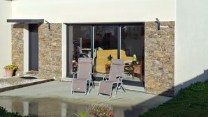A modern house with a stone facade, a large sliding glass door leading to an interior with a sofa and chairs, and a small outdoor seating area with two lounge chairs. - Ground Floor Holiday Home in Plestin des Greves (Plestin-les-Greves)