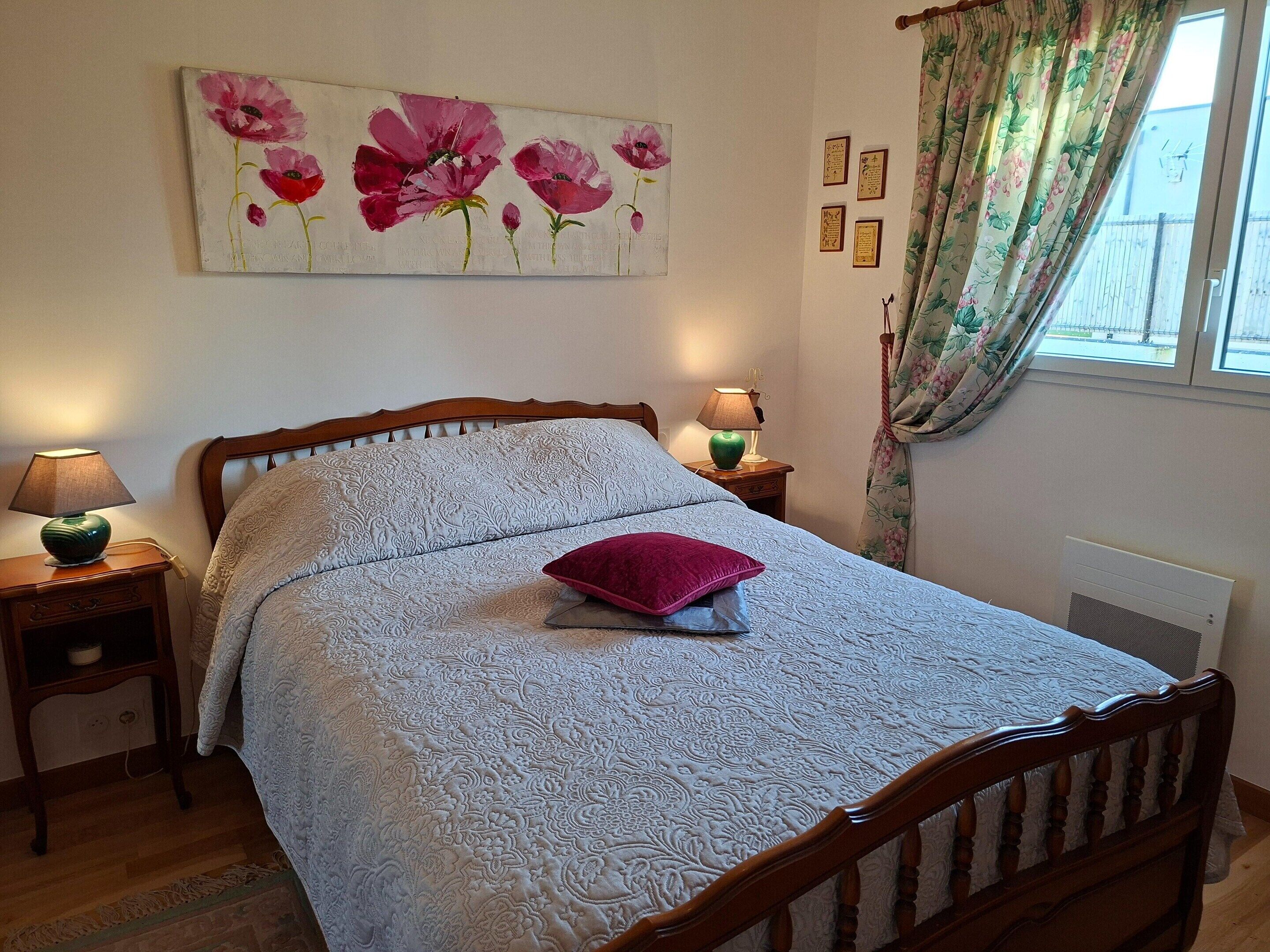 A bedroom with a bed, two bedside tables, a lamp, a floral painting, and a window with curtains.