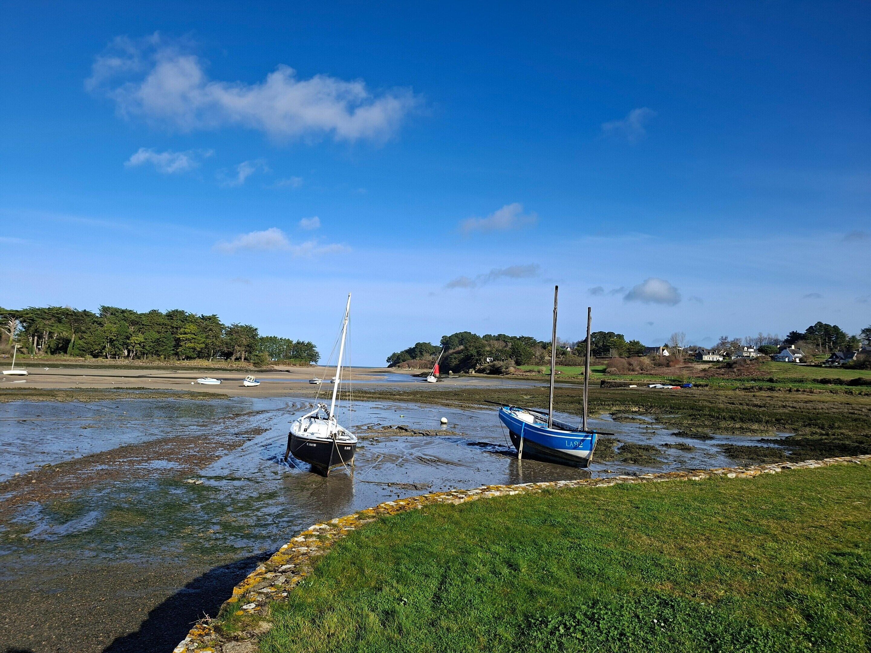 A coastal scene with boats anchored in shallow water, surrounded by grassy areas and distant houses.