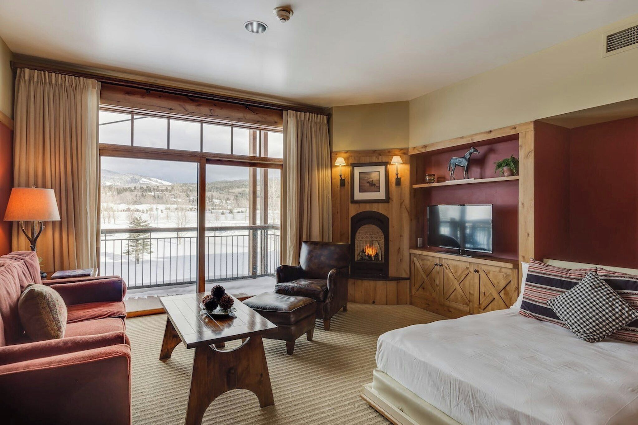 A hotel room with a large window, a fireplace, a TV, a sofa, and a bed.
