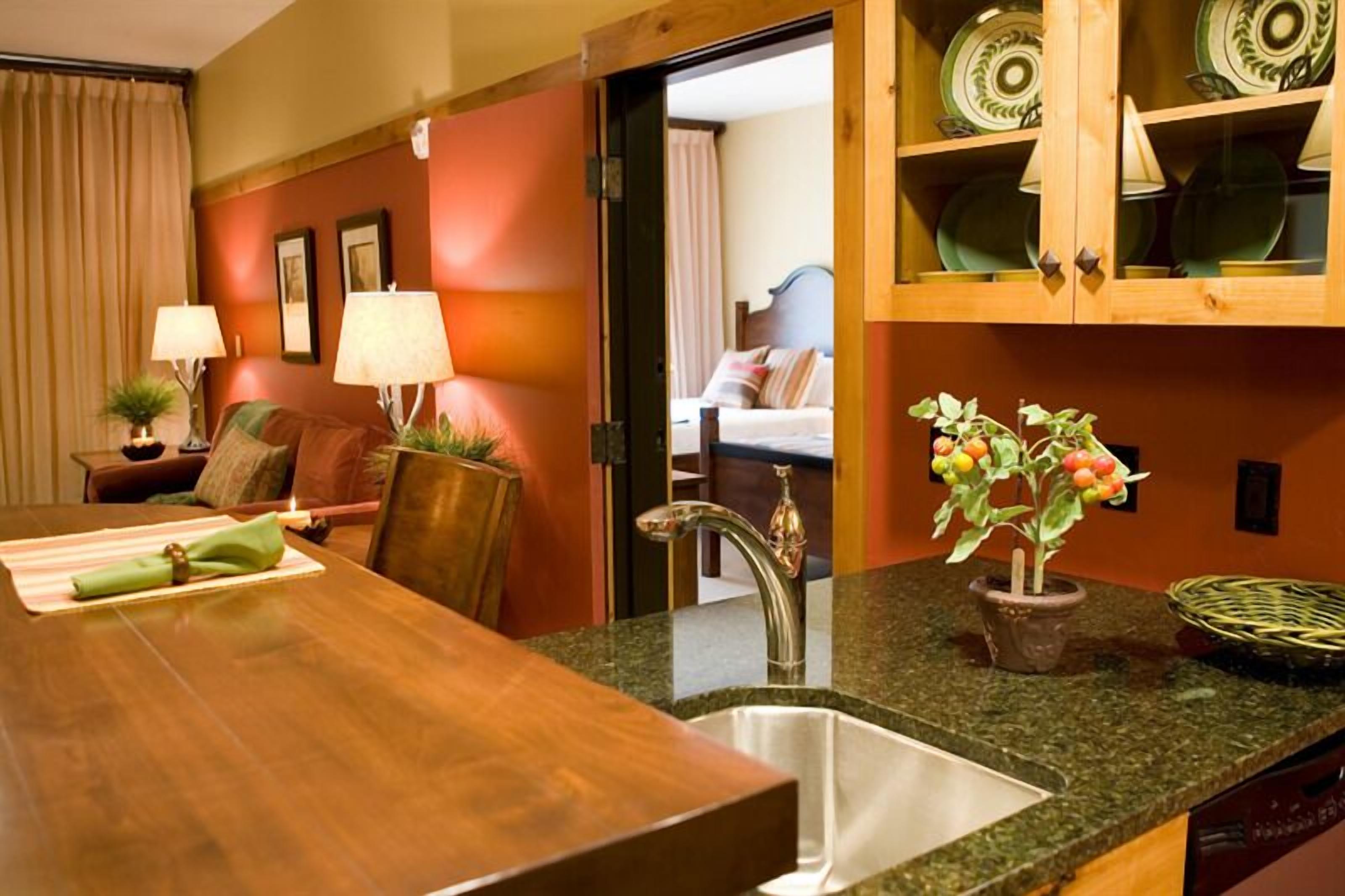 A hotel room with a wooden dining table, a sink, a potted plant, and a cabinet with plates.
