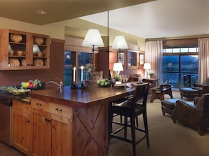 A spacious kitchen with wooden cabinets, a large island, and a dining area with chairs. There are two windows with curtains, and a balcony with a view. - Teton Springs Lodge And Spa (Victor)