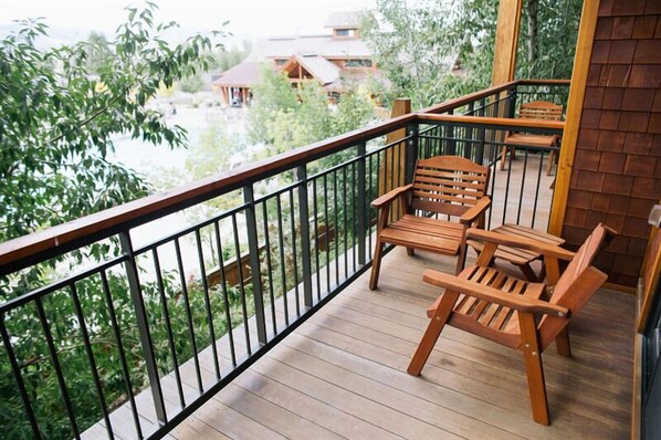 Una terraza de madera con barandilla, dos sillas de madera y vistas a edificios y árboles al fondo.