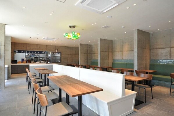 A modern restaurant with long wooden tables and cushioned seating, a kitchen area in the background, and a ceiling with recessed lighting. - The Breakfast Hotel Porto Ishigaki (Ishigaki)