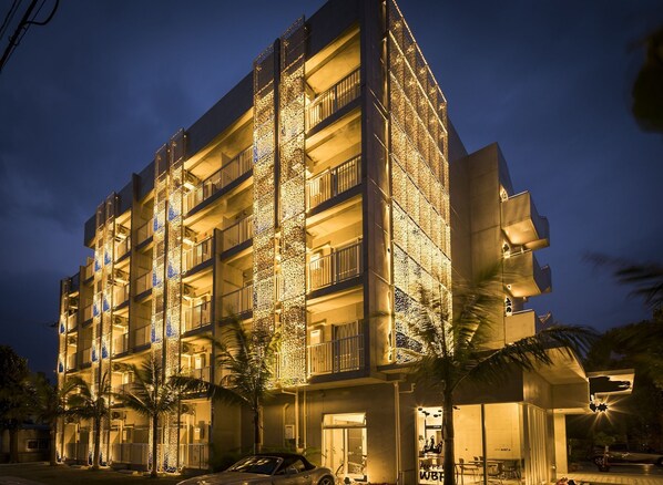 Un hotel de varios pisos con balcones, palmeras y un coche estacionado afuera.