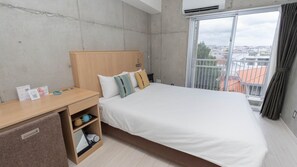 Quarto de hotel com cama, mesa de cabeceira de madeira, pequena estante com objetos e janela com vista para edifícios.