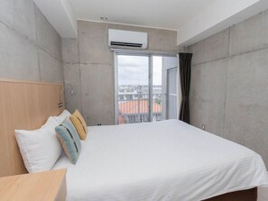 A hotel room with a bed, a wooden headboard, a window with a view of buildings, and an air conditioning unit. - The Breakfast Hotel Porto Ishigaki (Ishigaki)