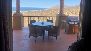 En balkon med et rundt bord og stole, grill og udsigt over ocean og bakker.