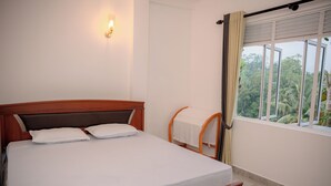 Ein ordentlich bezogenes Bett mit weißen Leinen und Kissen, ein hölzerner Kopfteil, ein kleiner Holzstuhl und ein Fenster mit Vorhängen.
