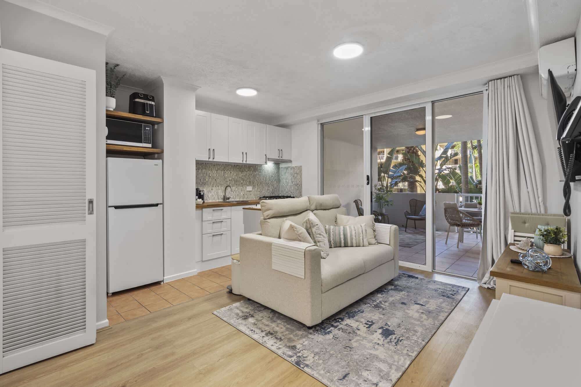 A modern kitchen with white cabinets, a microwave, and a refrigerator. There is a beige sofa with pillows in the living area, and a sliding glass door leading to a balcony with outdoor furniture.