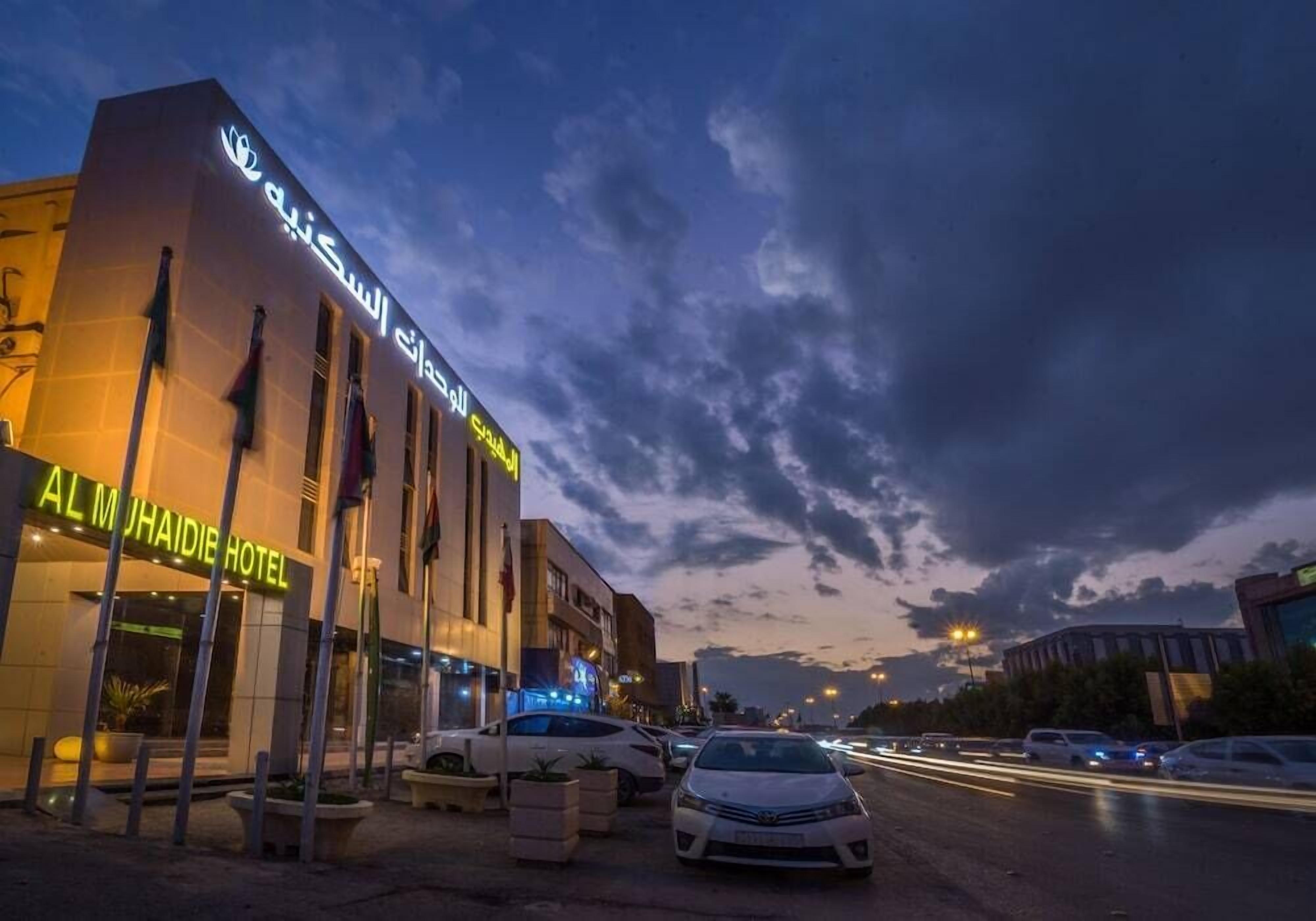 A hotel with a modern facade, a neon sign, and a parking lot with several cars.