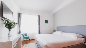 A modern hotel room with a bed, a desk, a chair, a TV, and a window with curtains.