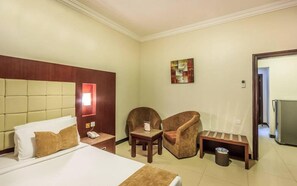 Habitación de hotel con cama, dos sillas, una mesita, un cesto de basura y un cuadro en la pared.