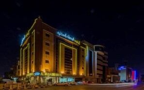 Hotel com placa em árabe bem visível, fachada moderna e estacionamento com vários carros.