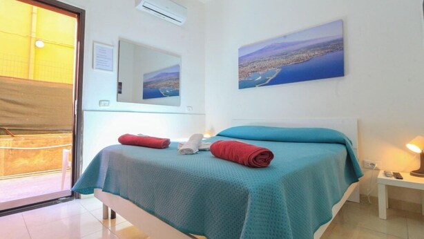 A bedroom with a bed, a bedside table with a lamp, a window with a view of a cityscape, and a door leading to an outdoor area. - Catania Aeroporto (Catania)