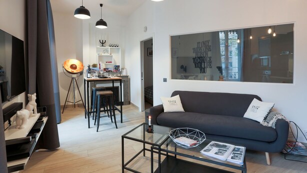 A modern living room with a sofa, a coffee table, and a TV. There is a dining area with a table and chairs, and a large window with a view of the city. - Suite Garibaldi Cosy Design Lyon Centre (Lyon)