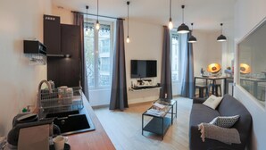 A modern living room with a sofa, coffee table, and TV. There is a kitchen area with a fridge and a coffee machine. The room has large windows with curtains. - Suite Garibaldi Cosy Design Lyon Centre (Lyon)