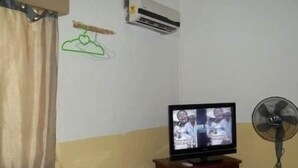 A room with a television on a wooden stand, a white air conditioner on the wall, a fan, and a small table with a white appliance. - Haoxin Hotel (Accra)
