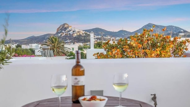A table with a bottle and glasses, a bowl of food, and a view of mountains and buildings. - Anemonisia (Skiros)