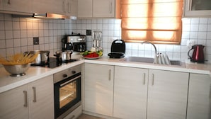 A modern kitchen with white cabinets, a stove, a coffee maker, and a window with blinds. - Anemonisia (Skiros)