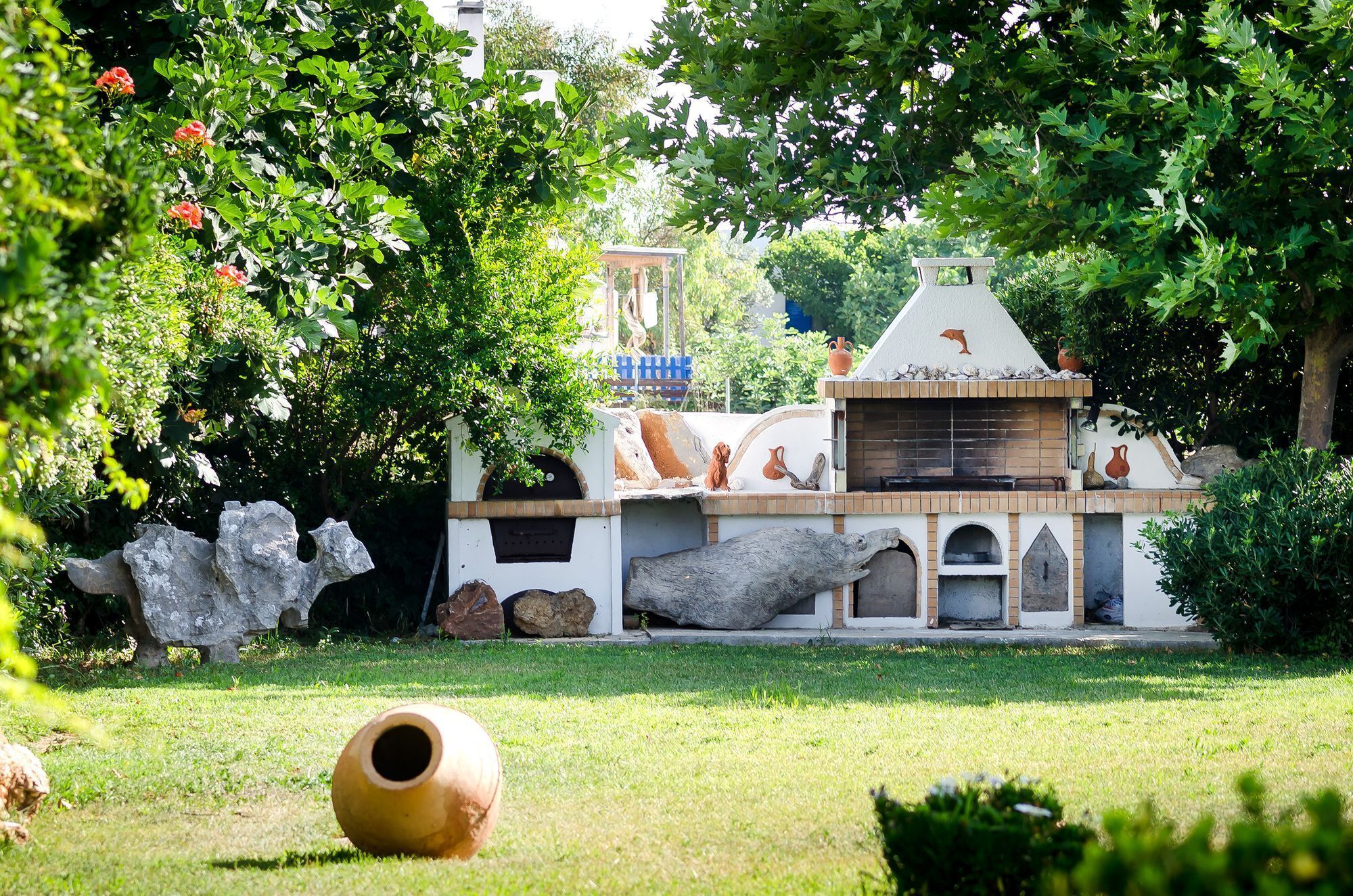 A rustic outdoor kitchen with a stone fireplace, surrounded by lush greenery and a large rock sculpture.