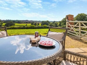 A round table with a coffee pot, cups, and a plate of food, surrounded by wicker chairs, set outdoors with a view of green fields and a brick structure in the background.
