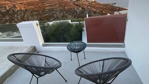 Ein Balkon mit zwei schwarzen Stuhlen aus Draht und einem kleinen runden Tisch, mit Blick auf eine Küstenlandschaft mit Hügeln und einem entfernten Meerblick.