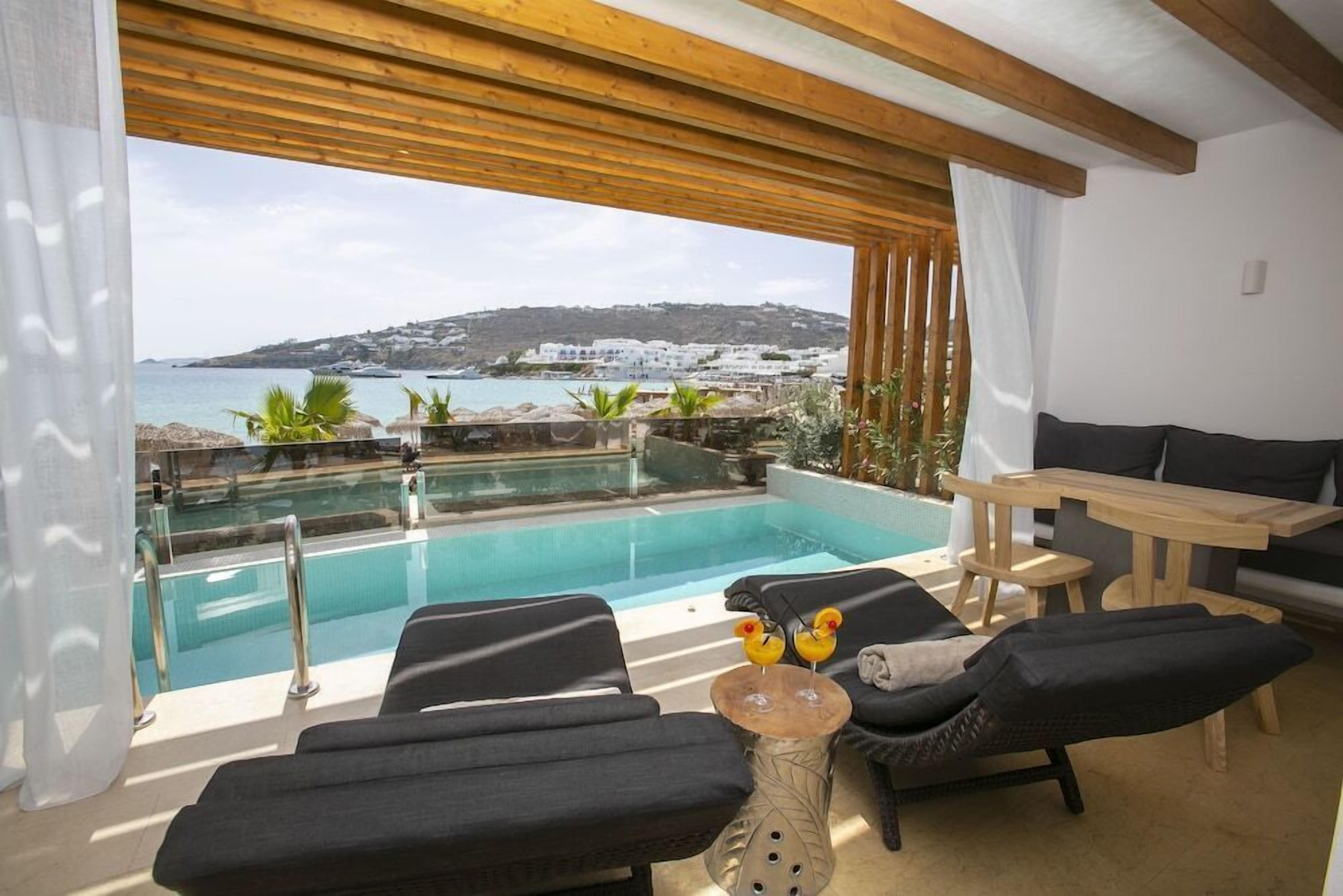 A sunlit outdoor seating area with a pool, lounge chairs, and a table, overlooking a coastal landscape with buildings and hills.