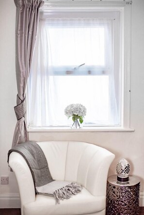 Una poltrona bianca con una coperta grigia, un tavolino di metallo decorativo, una finestra con tende leggere e un vaso con fiori bianchi.