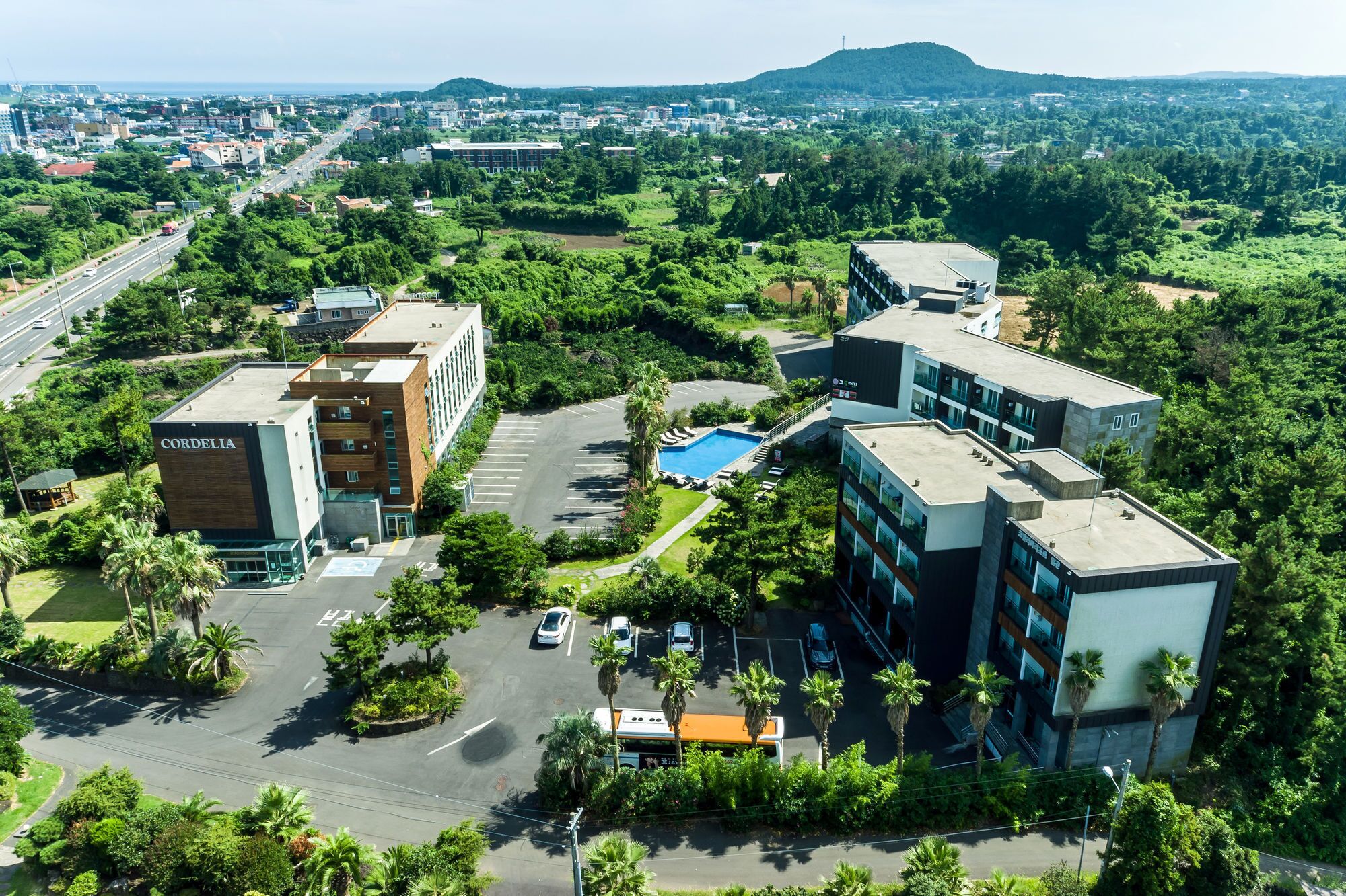 An aerial view of a modern hotel complex with multiple buildings, a swimming pool, and surrounding greenery.
