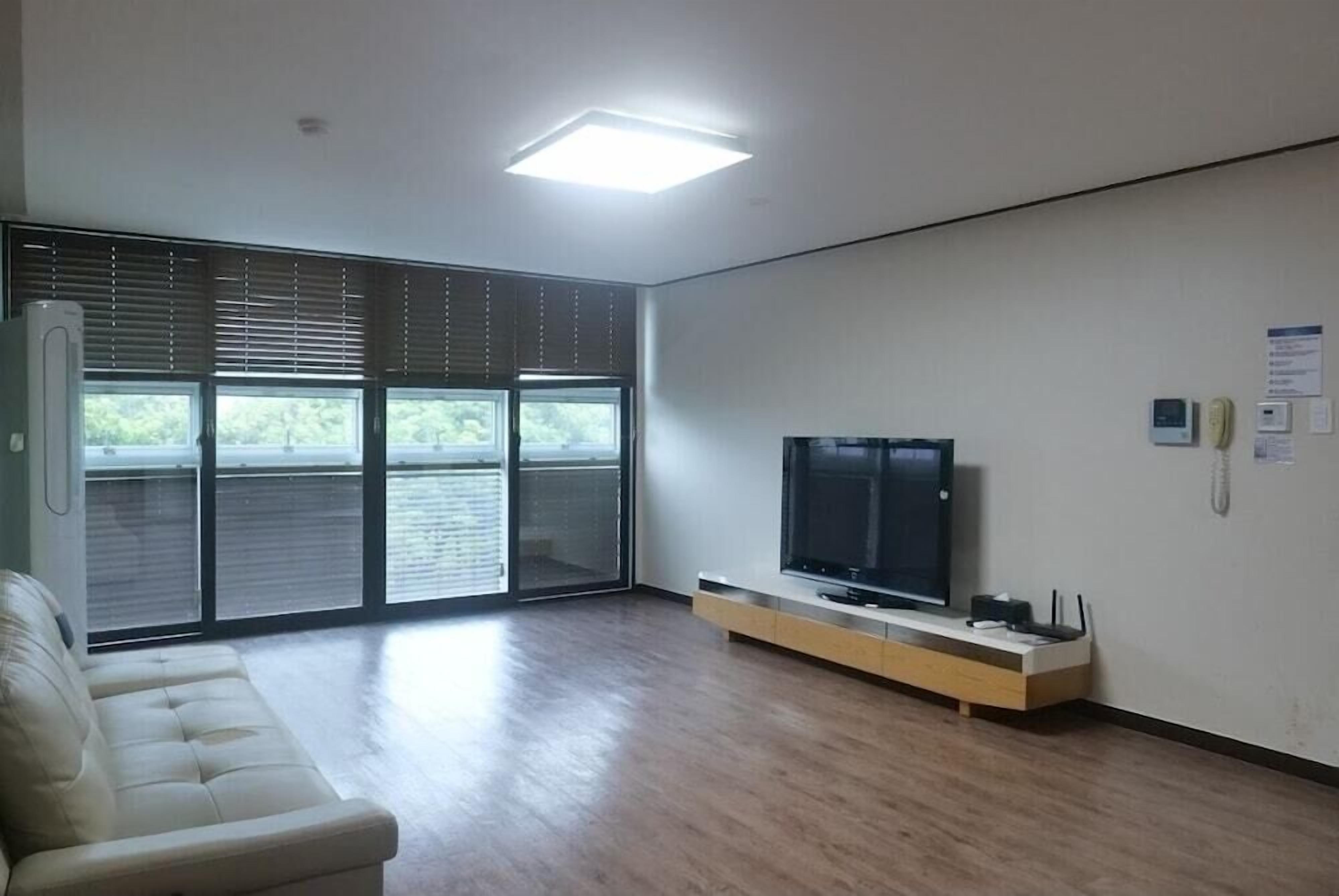 Een ruime woonkamer met een grote flatscreen televisie op een houten standaard, een schuifpui naar een balkon en een witte bank.