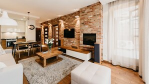 A modern living room with a brick wall, a flat-screen TV, a fireplace, and a dining area with a kitchen in the background. - Exclusive Apartments (Kraków)