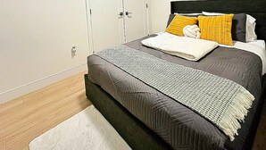 A bedroom with a bed, a nightstand, a window with blinds, a door, and a wall-mounted picture. - Luxury 2-Bedroom Suite at Bear Mountain | Victoria (Langford)