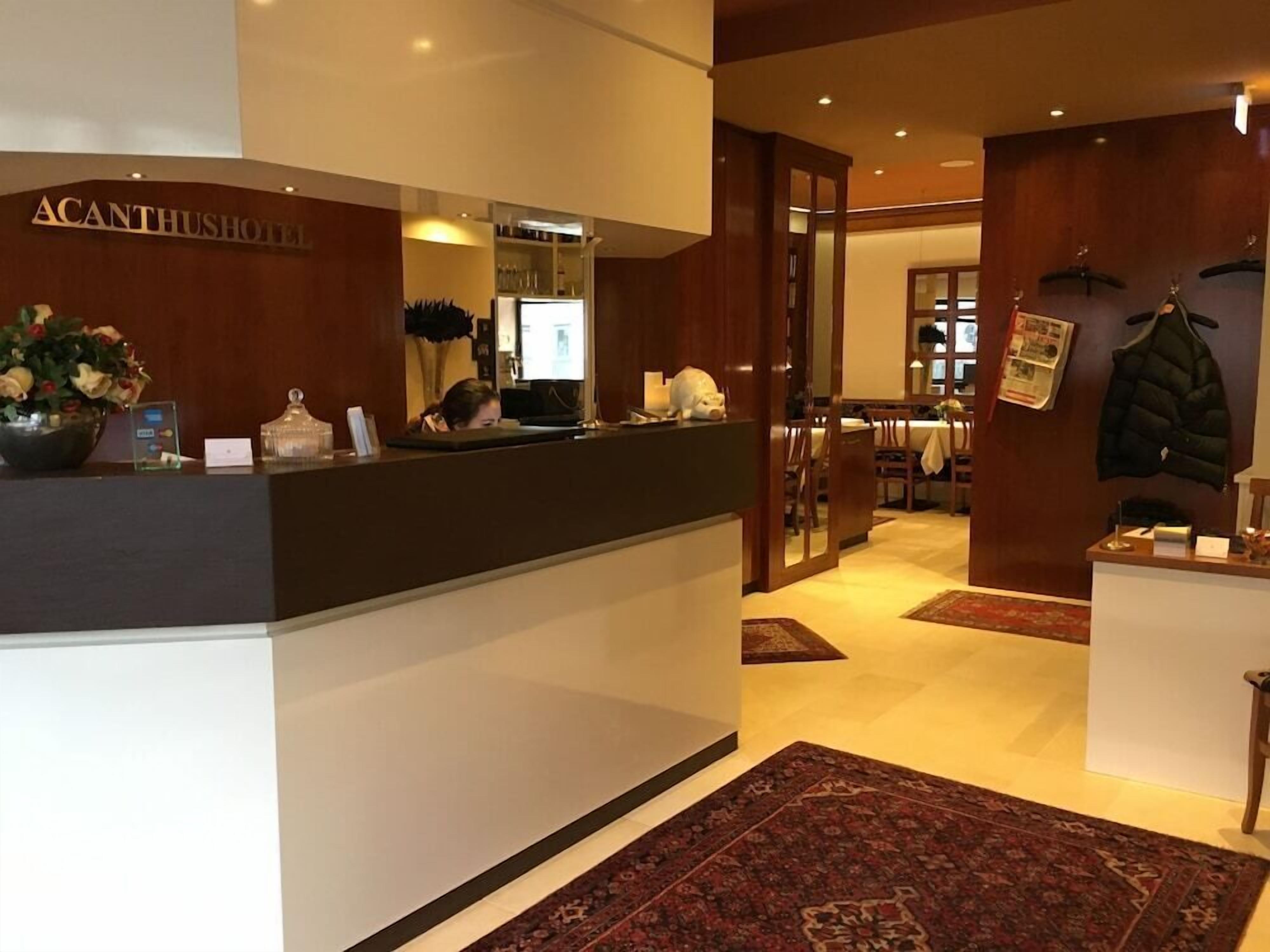 Ein Hotel-Empfangsbereich mit Empfangstresen, Spiegel, Garderobe und einem Essbereich im Hintergrund.
