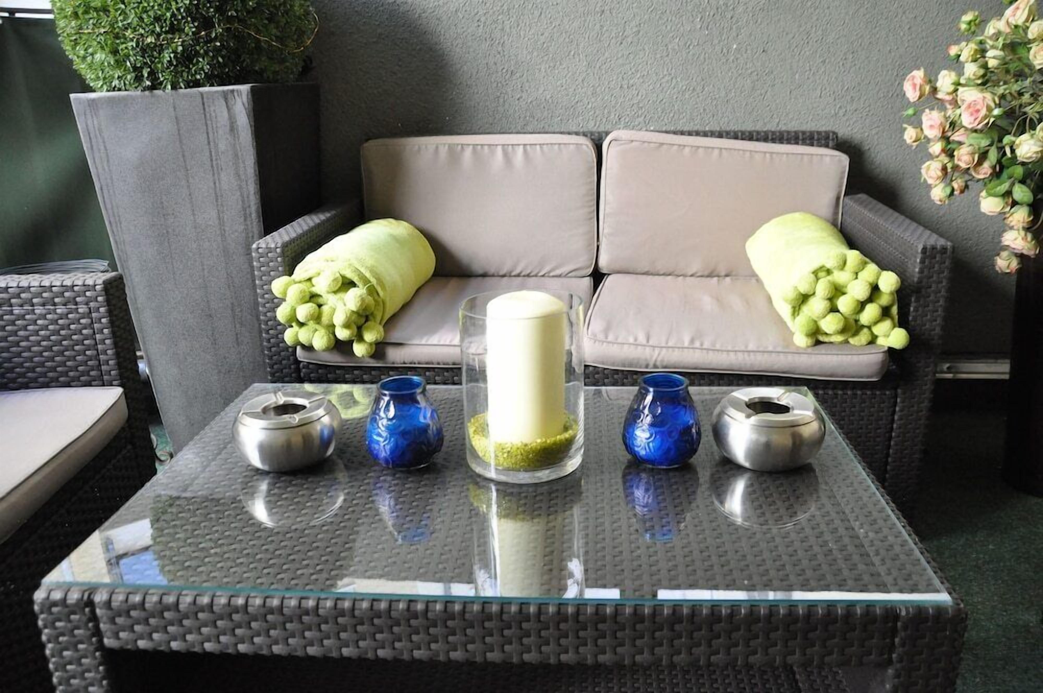 Ein Tisch mit Glasplatte, eine weiße Kerze und blaue Vasen, ein graues Sofa mit grünen Kissen und eine Topfpflanze im Hintergrund.
