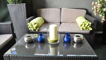 Ein Tisch mit Glasplatte, eine weiße Kerze und blaue Vasen, ein graues Sofa mit grünen Kissen und eine Topfpflanze im Hintergrund.