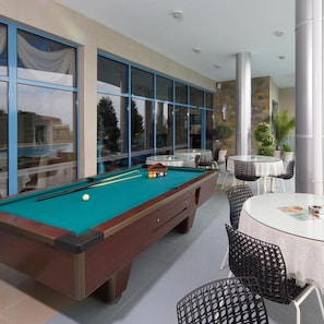 A pool table with a green felt surface, surrounded by black chairs and a white table with a lace tablecloth. - Blue Bay Hotel (Nessebar)