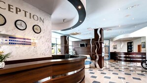 A reception area with a curved wooden reception desk, a wall clock, and a digital display. - Blue Bay Hotel (Nessebar)
