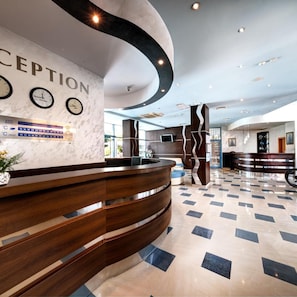 A reception area with a curved wooden reception desk, a wall clock, and a digital display. - Blue Bay Hotel (Nessebar)