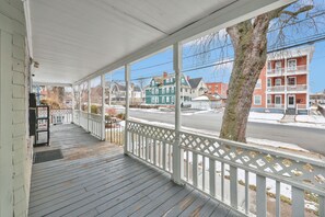 A covered porch with a white railing, overlooking a street with multi-story buildings and a tree.