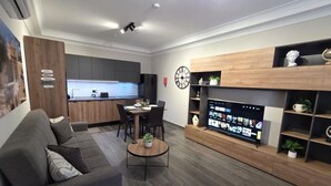 A modern living room with a sofa, coffee table, TV, and a kitchen in the background. - Cosy Ground FL APT - Msida Marina Area (Msida)
