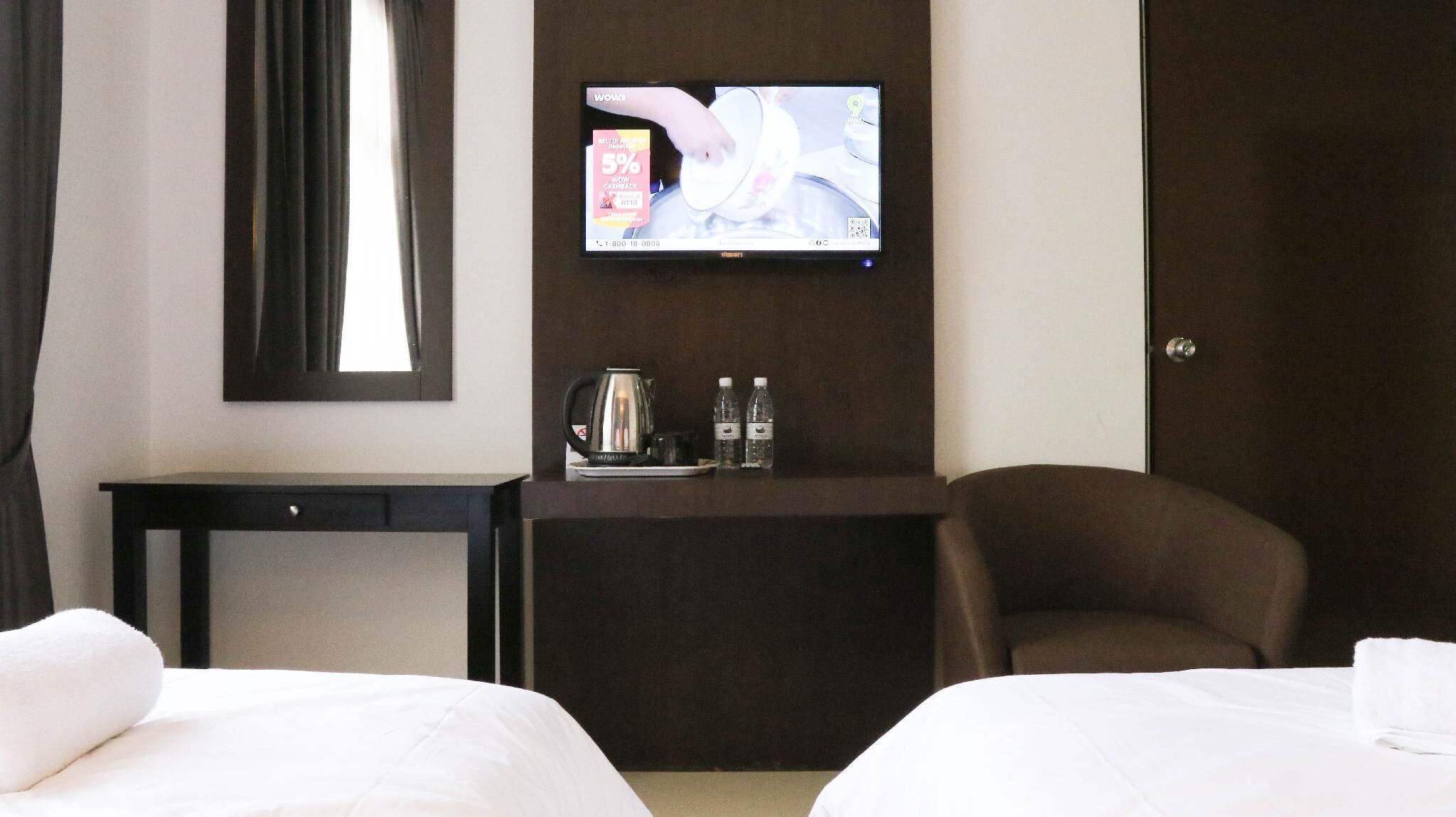 Ein Hotelzimmer mit zwei Betten, einem an der Wand befestigten Fernseher, einem kleinen Tisch mit Kaffeemaschine und Wasserflaschen sowie einem Sessel.
