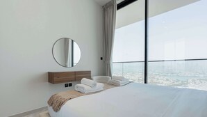 Ein modernes Hotelzimmer mit einem großen Bett, einem runden Spiegel, einem Nachttisch aus Holz und Blick auf eine Stadtlandschaft durch große Fenster.