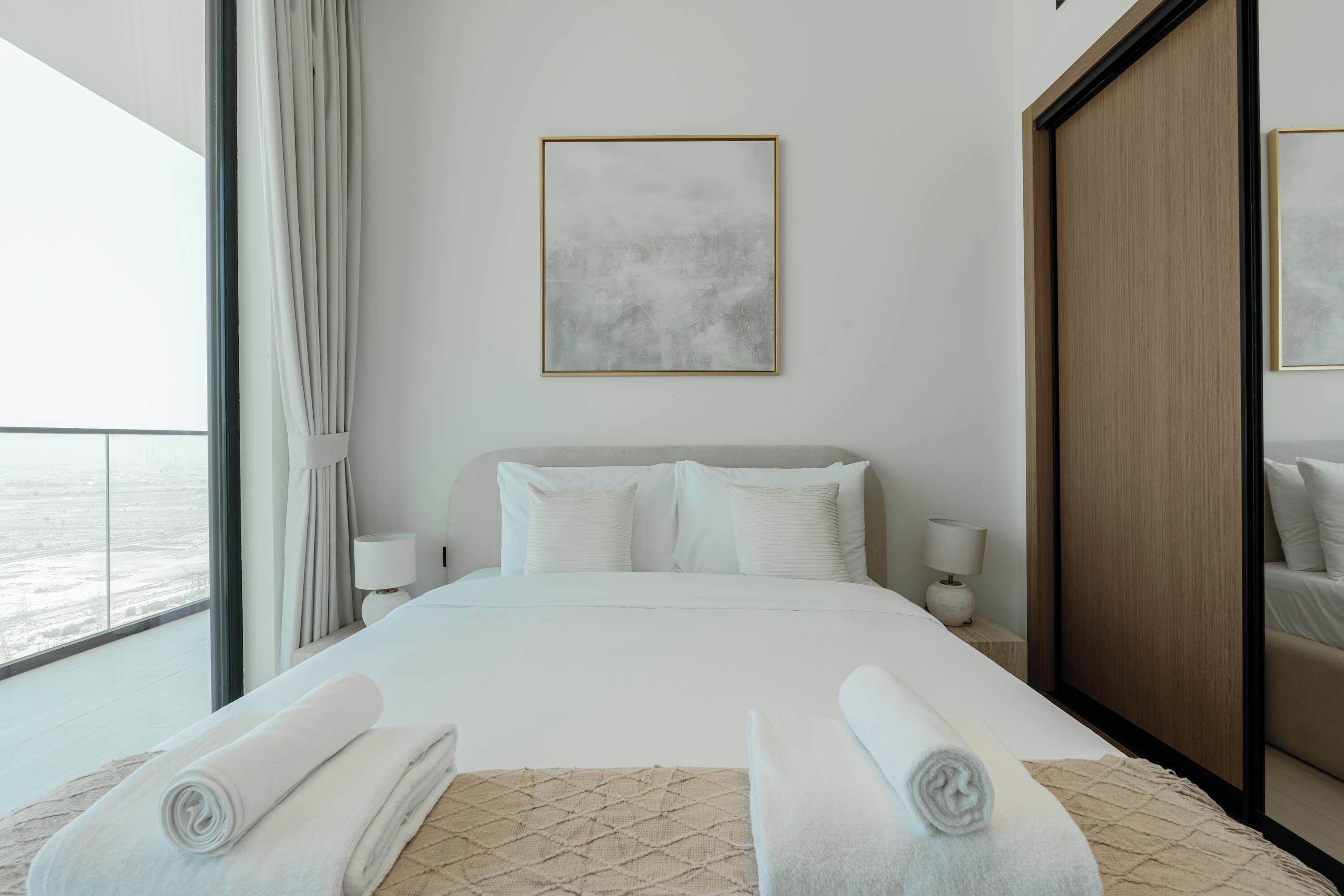 A modern hotel room with a large bed, a bedside table with a lamp, a view of the sea, and a framed abstract painting on the wall.