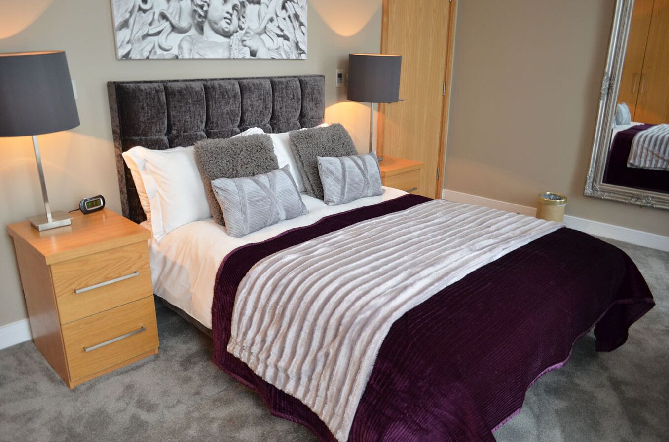 A neatly made bed with a headboard, two bedside tables with lamps, a drawer unit, and a wall-mounted artwork.