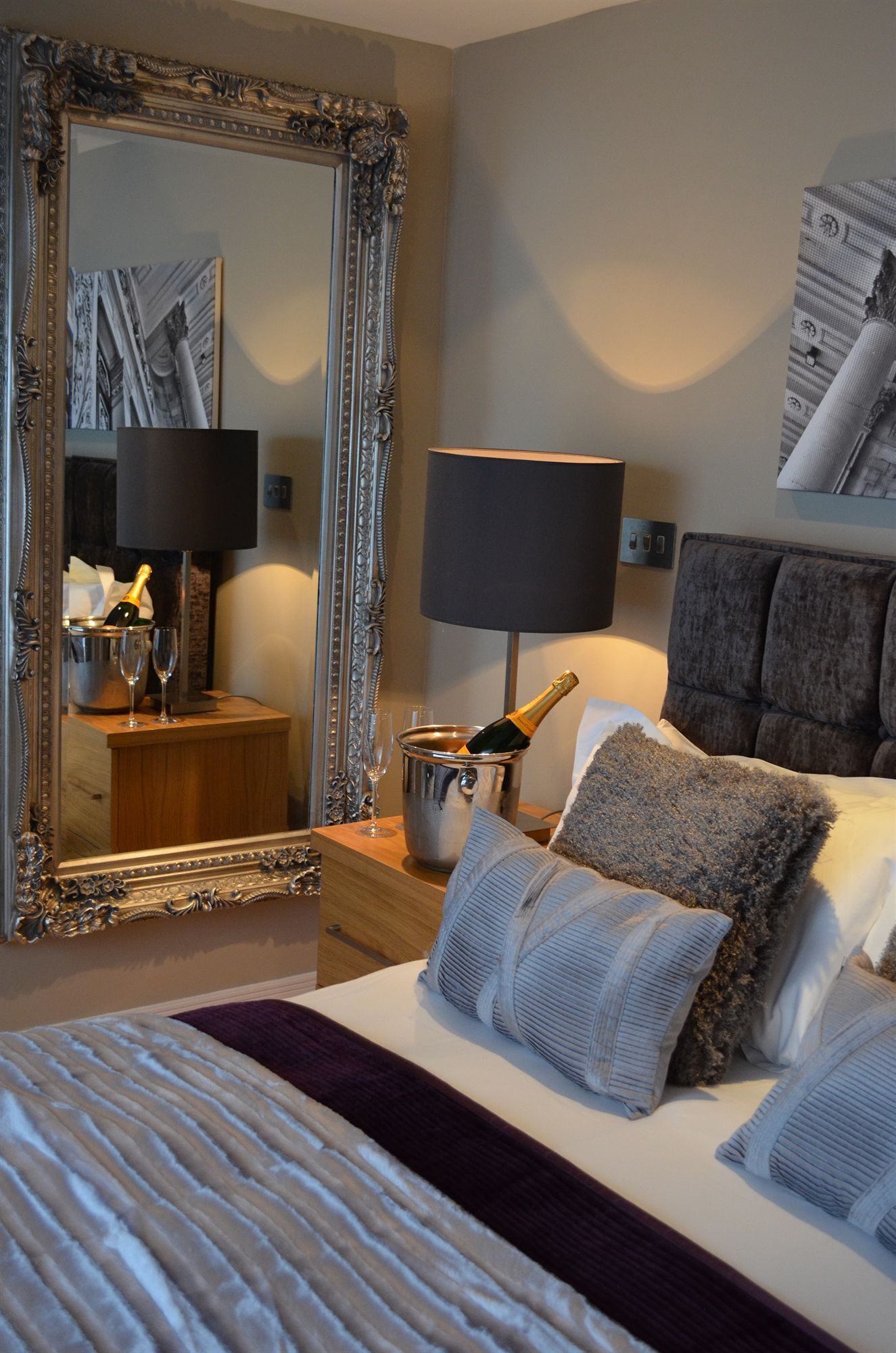 Une chambre d’hôtel avec un grand miroir décoratif, un lit recouvert de plusieurs coussins, une table de chevet sur laquelle se trouvent une bouteille de champagne et des verres, une lampe et un tableau encadré accroché au mur.