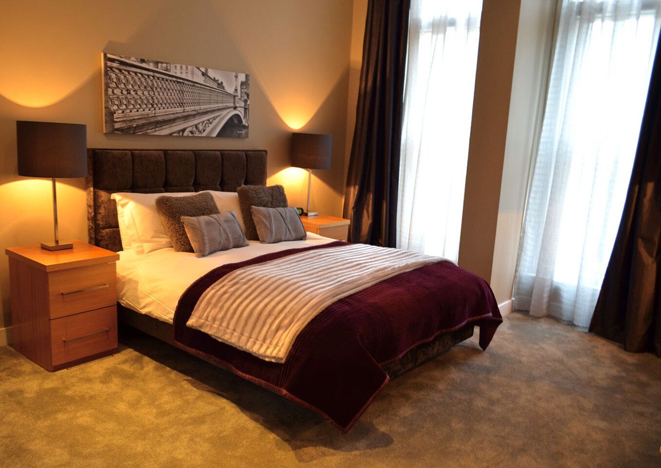 A hotel room with a large bed, two bedside tables, a headboard, a wall-mounted artwork, and a window with curtains.