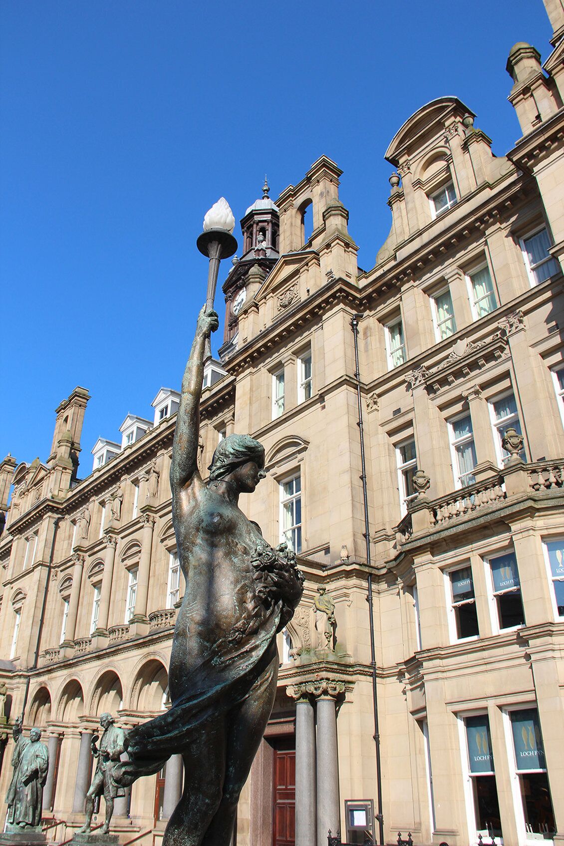 A statue of a woman holding a lamp in front of a large, ornate building with classical architecture.