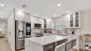 A modern kitchen with white cabinetry, stainless steel appliances, and a marble island with bar stools. - IRB Cypress & Sun (Indian Rocks Beach)